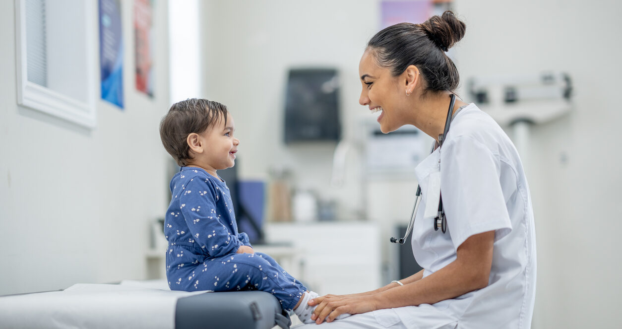 A sweet little toddler sits up on an exam table in a doctors office as her female doctor conducts a routine check-up. The little girl is dressed casually and appears happy as her doctor engages with her.