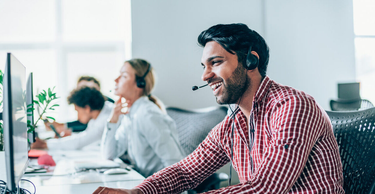 Smiling handsome ethnicity businessman working in call center. Shot of a cheerful young man working in a call center with his team. Confident male operator is working with colleagues. Call center operators sitting in a row at desks.