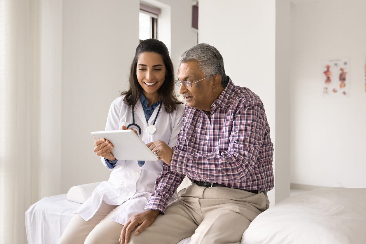 Excited old Indian patient man and happy young doctor woman using digital tablet together, looking at display, talking, discussing HbA1c medical test results, modern electronic diagnosing